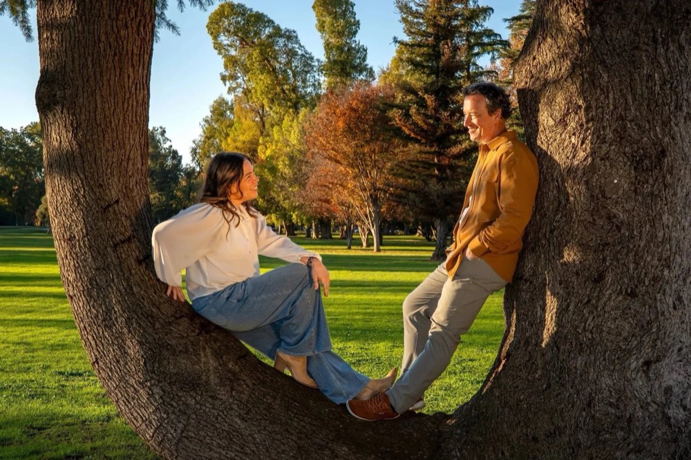 Molly and James Christensen smiling at each other while leaning against a large tree